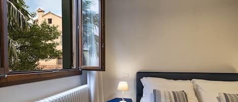 Una habitación de hotel con una cama, una ventana que ofrece vistas a edificios y árboles, un radiador y una pequeña mesa con una lámpara.