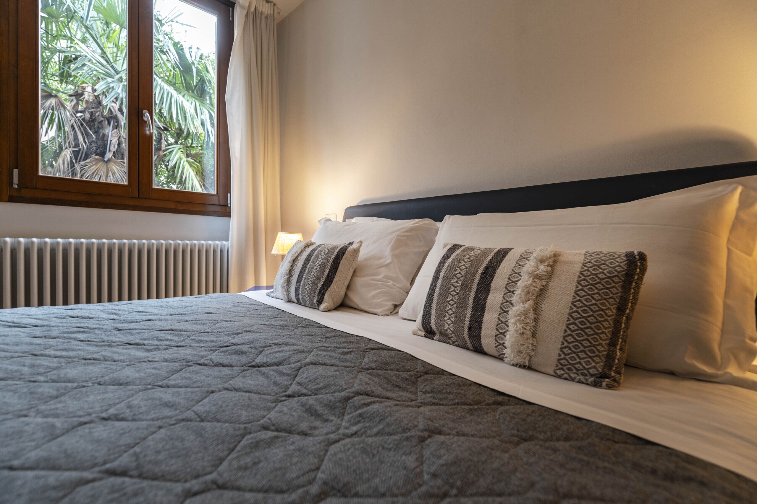 A neatly made bed with a quilted grey bedspread and patterned pillows, a window with a view of palm trees, and a bedside lamp.