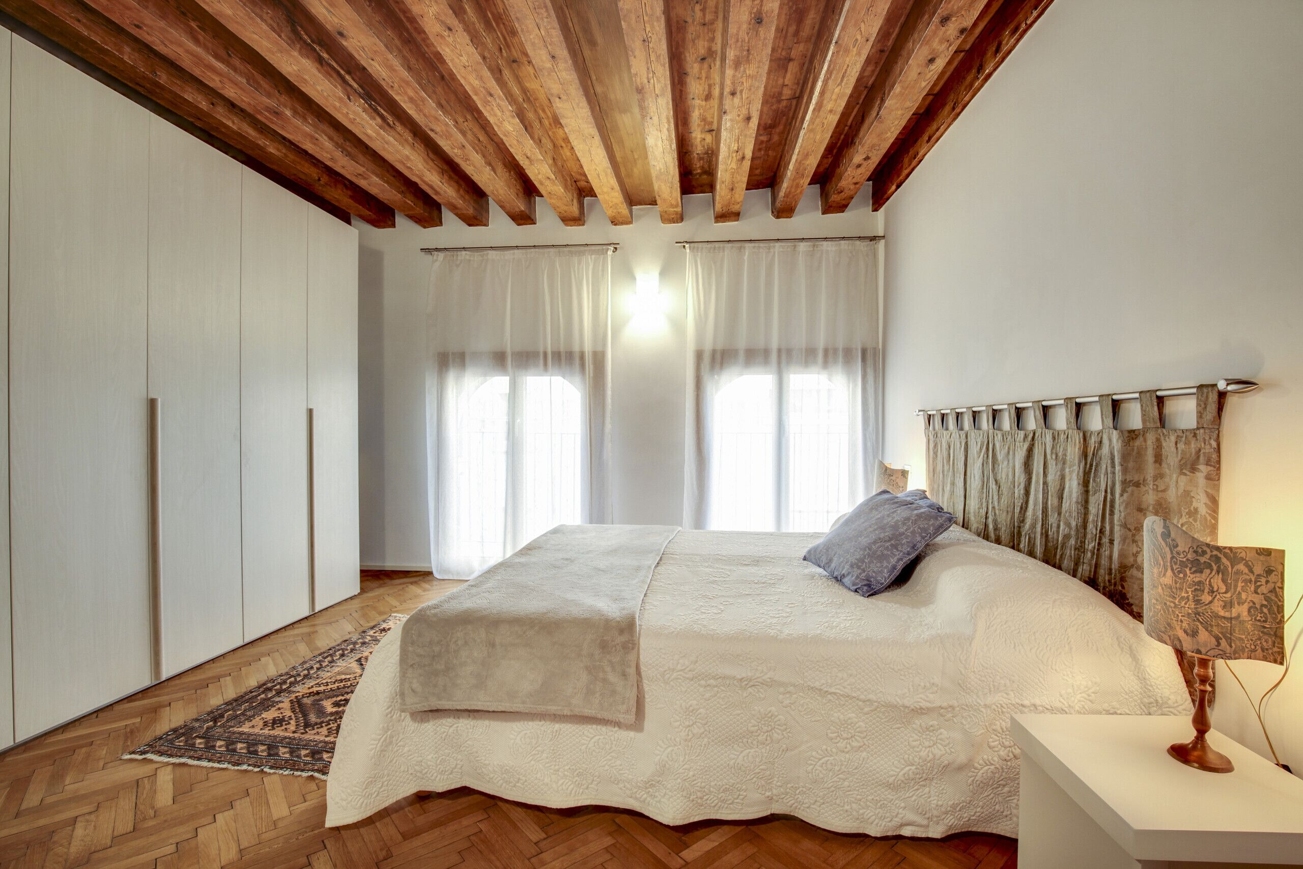 Een slaapkamer met een houten plafond, een bed met een witte dekbedovertrek, een kamerbreed tapijt met dessin en een nachtkastje met een lamp.