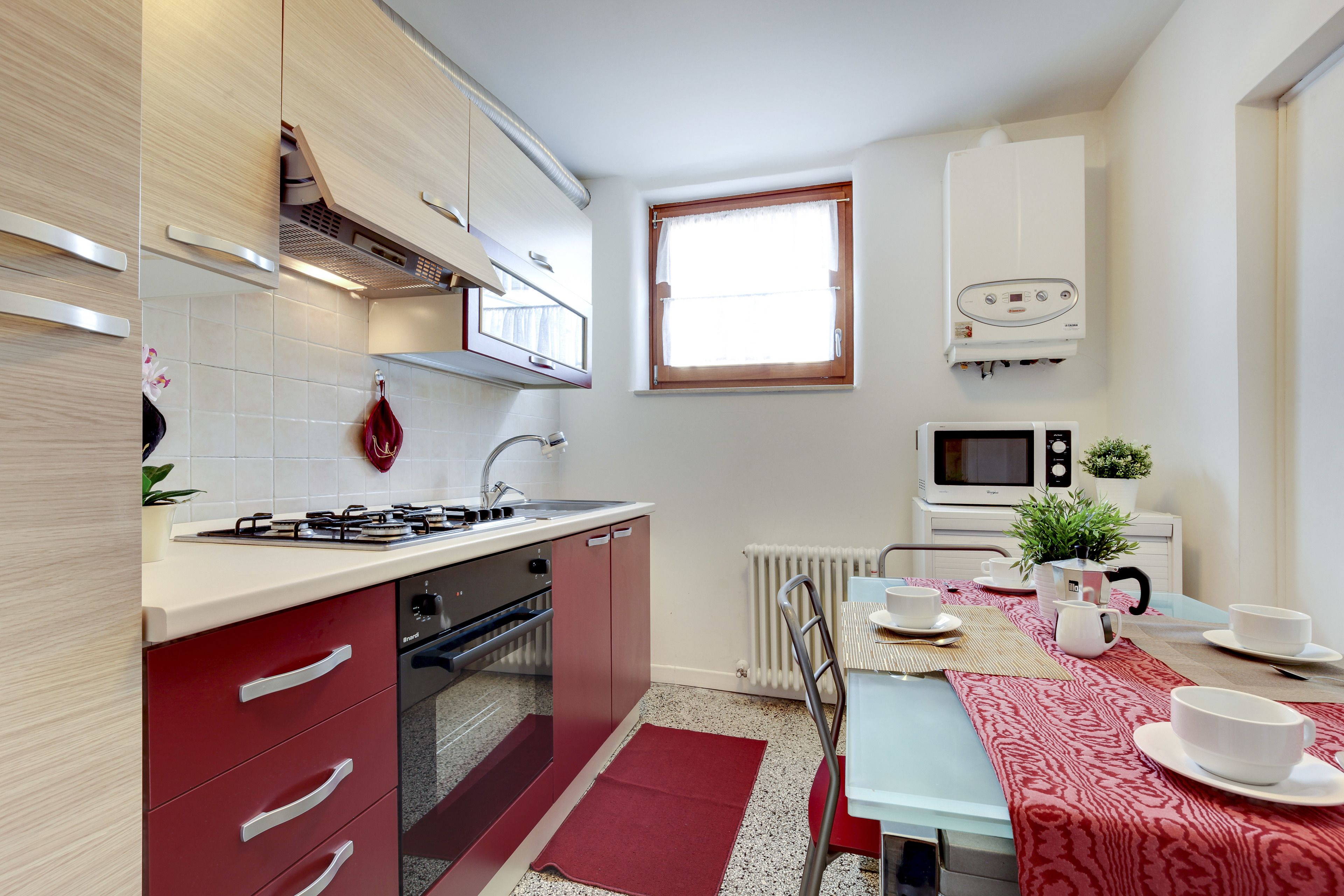 Une cuisine compacte équipée d’une cuisinière à gaz, d’un évier et d’un four à micro-ondes. On y trouve une table à manger avec un chemin de table rouge et des assiettes blanches.