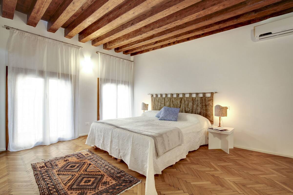 A bedroom with a wooden ceiling, a bed with a white bedspread, a patterned rug, and a nightstand with a lamp.