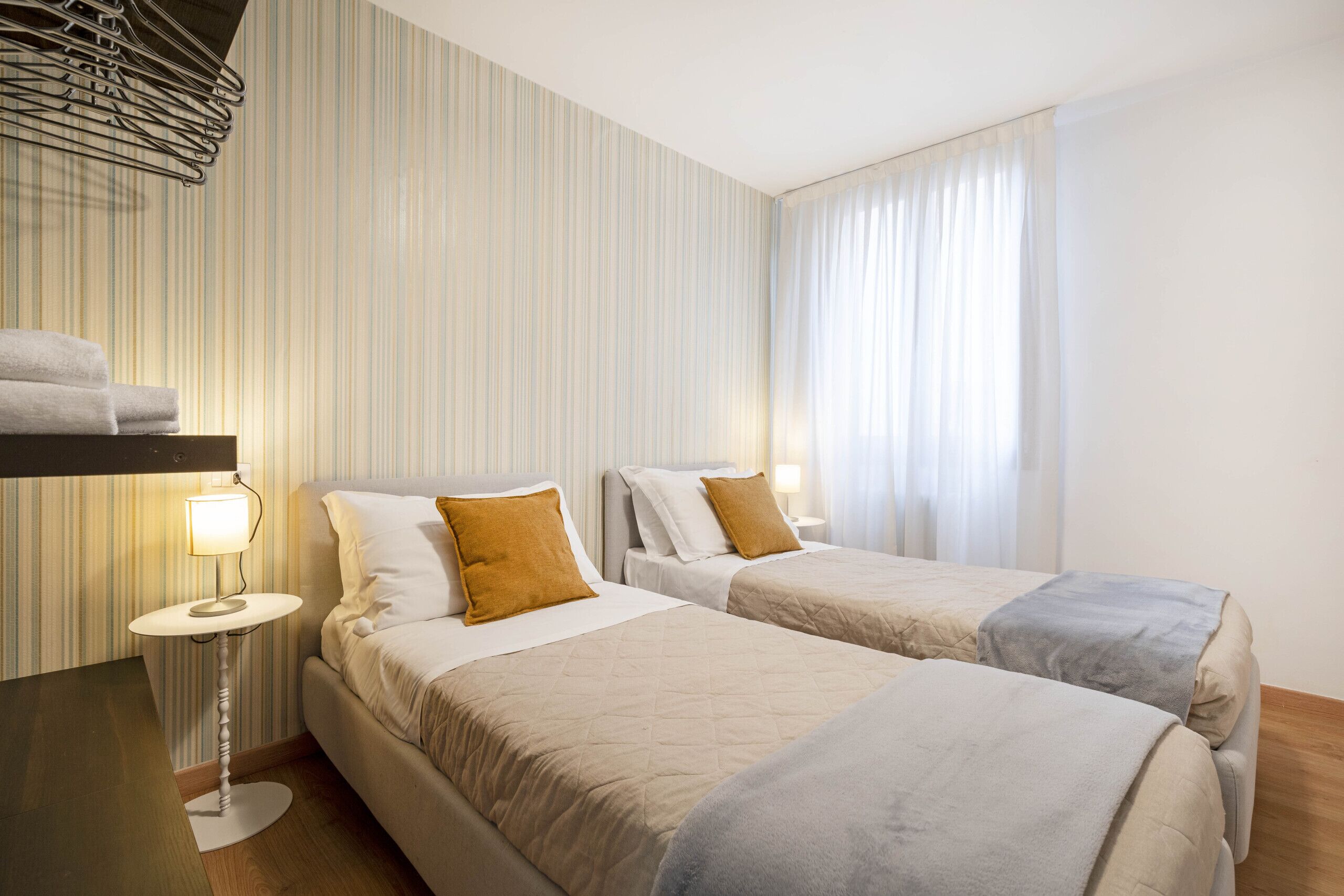 Habitación de hotel con dos camas, una mesita de noche con lámpara, papel tapiz a rayas y una ventana con cortinas.