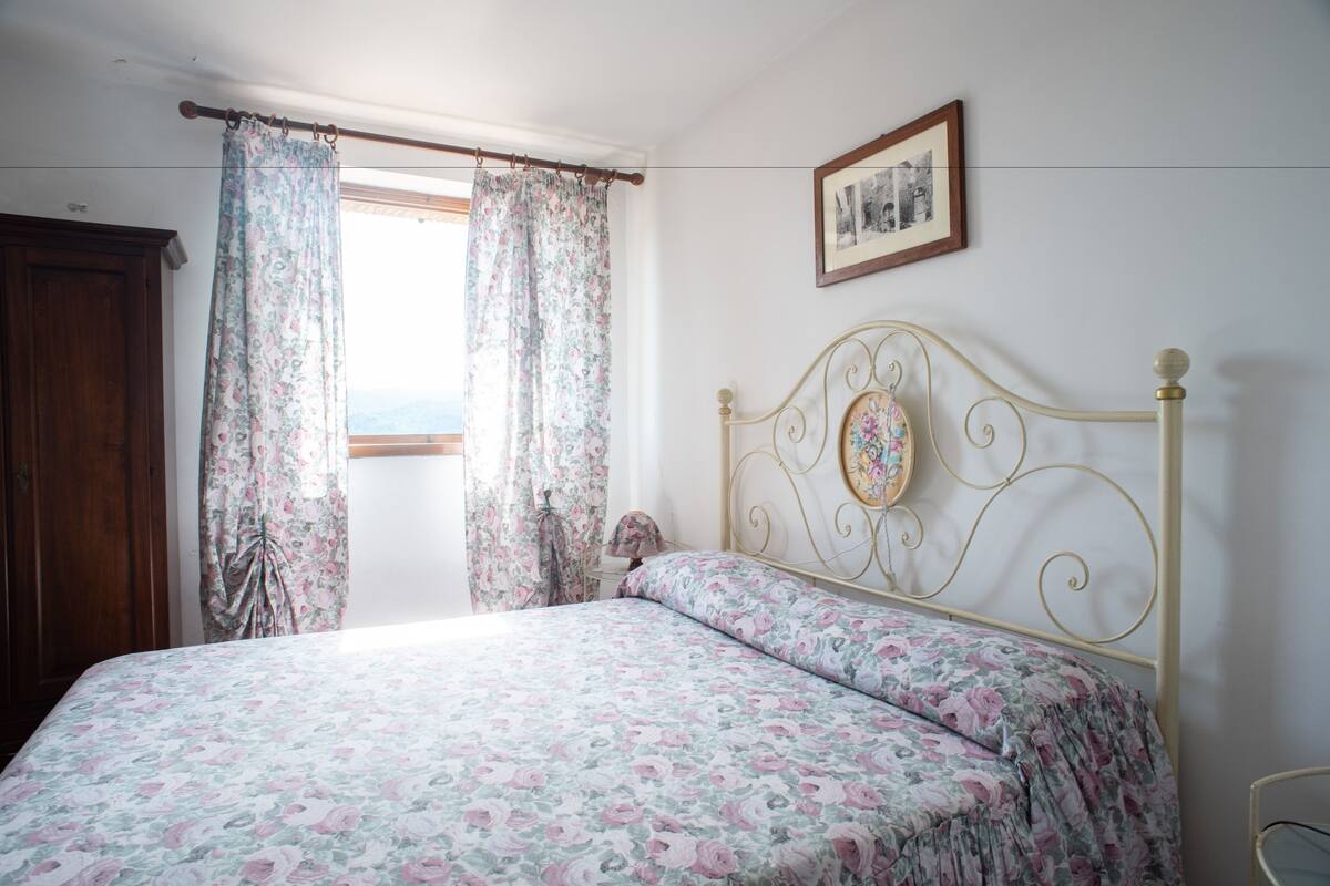 A bedroom with a floral bedspread, a wooden wardrobe, and a window with curtains.