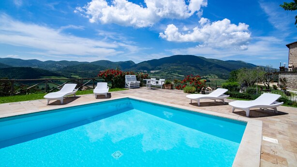 A swimming pool with lounge chairs and a table, surrounded by a stone deck, with a view of mountains and a clear sky. - Il Castello di Perchia (Spoleto)