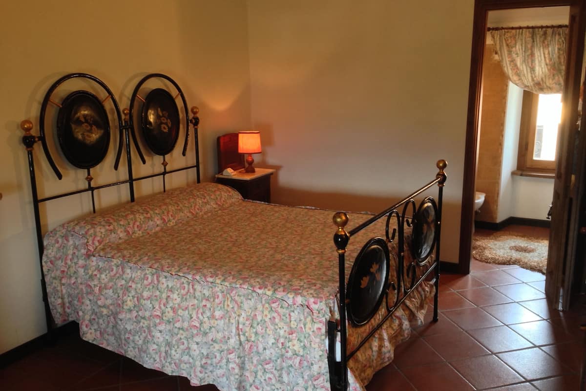 A bedroom with a bed, a nightstand with a lamp, and a door leading to another room.