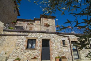 Un edificio de piedra con puerta negra, ventanas con contraventanas y balcones con plantas en macetas.