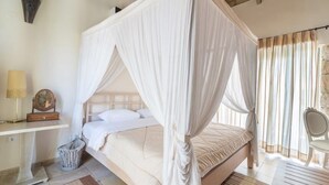 Un dormitorio con cama con dosel, una mesita de noche con lámpara, una silla y vistas al exterior a través de cortinas.