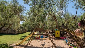 Un parque infantil con columpios, tobogán y zona de arena, rodeado de árboles y un muro de piedra.