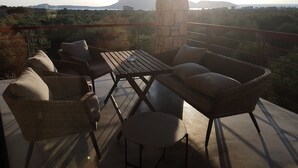 Un área de estar al aire libre techada con muebles de mimbre y una mesa, con vista a un paisaje pintoresco que incluye colinas y una masa de agua.