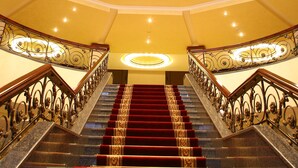 Una imponente escalera con barandales ornamentados y un tapete rojo, que conduce a una lámpara circular en el techo.