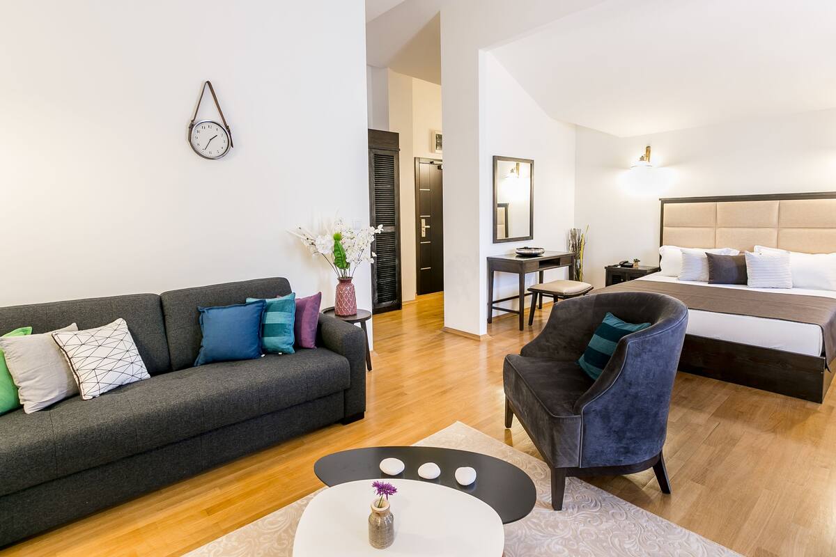 Ein modernes Wohnzimmer mit einem grauen Sofa, einem runden Couchtisch und einem blauen Sessel. Es gibt ein Bett mit Kopfteil sowie einen kleinen Schreibtisch mit Stuhl.
