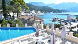 Piscina com espreguiçadeiras e guarda-sóis, vista de uma marina com barcos, e edifícios ao fundo.