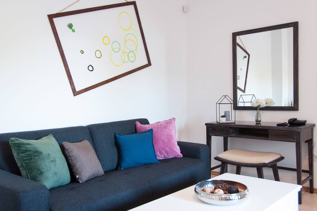 Ein modernes Wohnzimmer mit einem dunkelgrauen Sofa, mehreren Kissen, einem weißen Couchtisch mit dekorativer Schale, einem dunklen Holztisch und einem großen Spiegel.