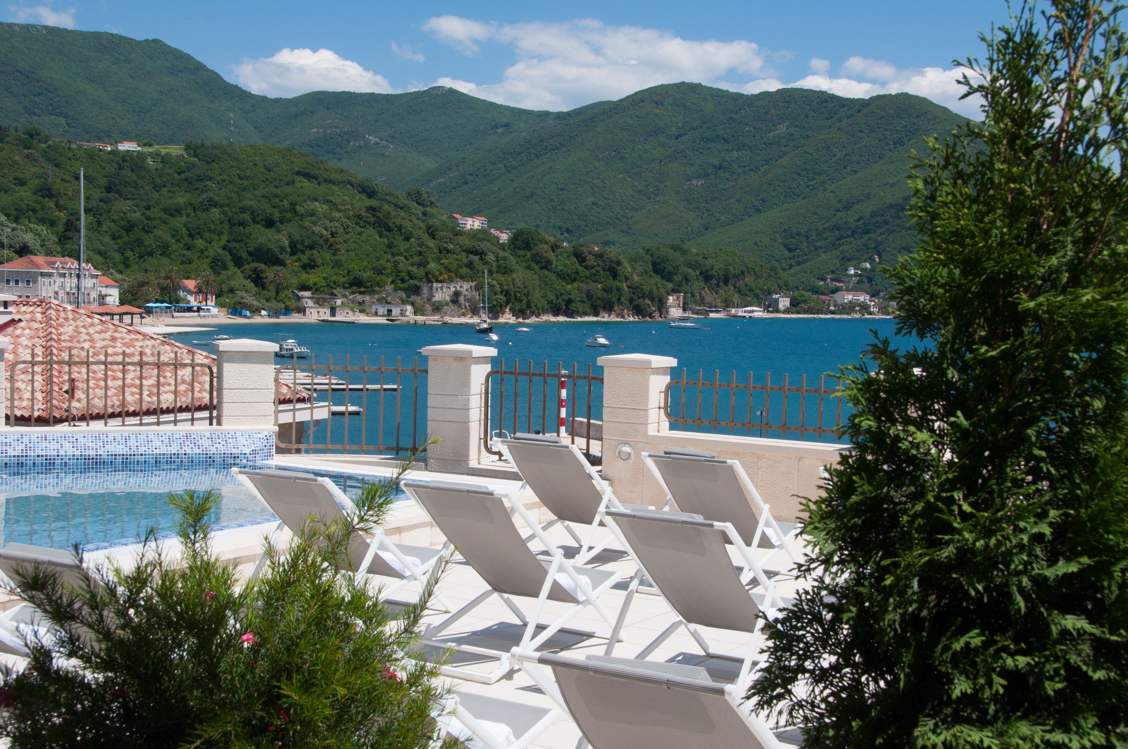 A pool area with lounge chairs, a pool, and a view of the sea and mountains.
