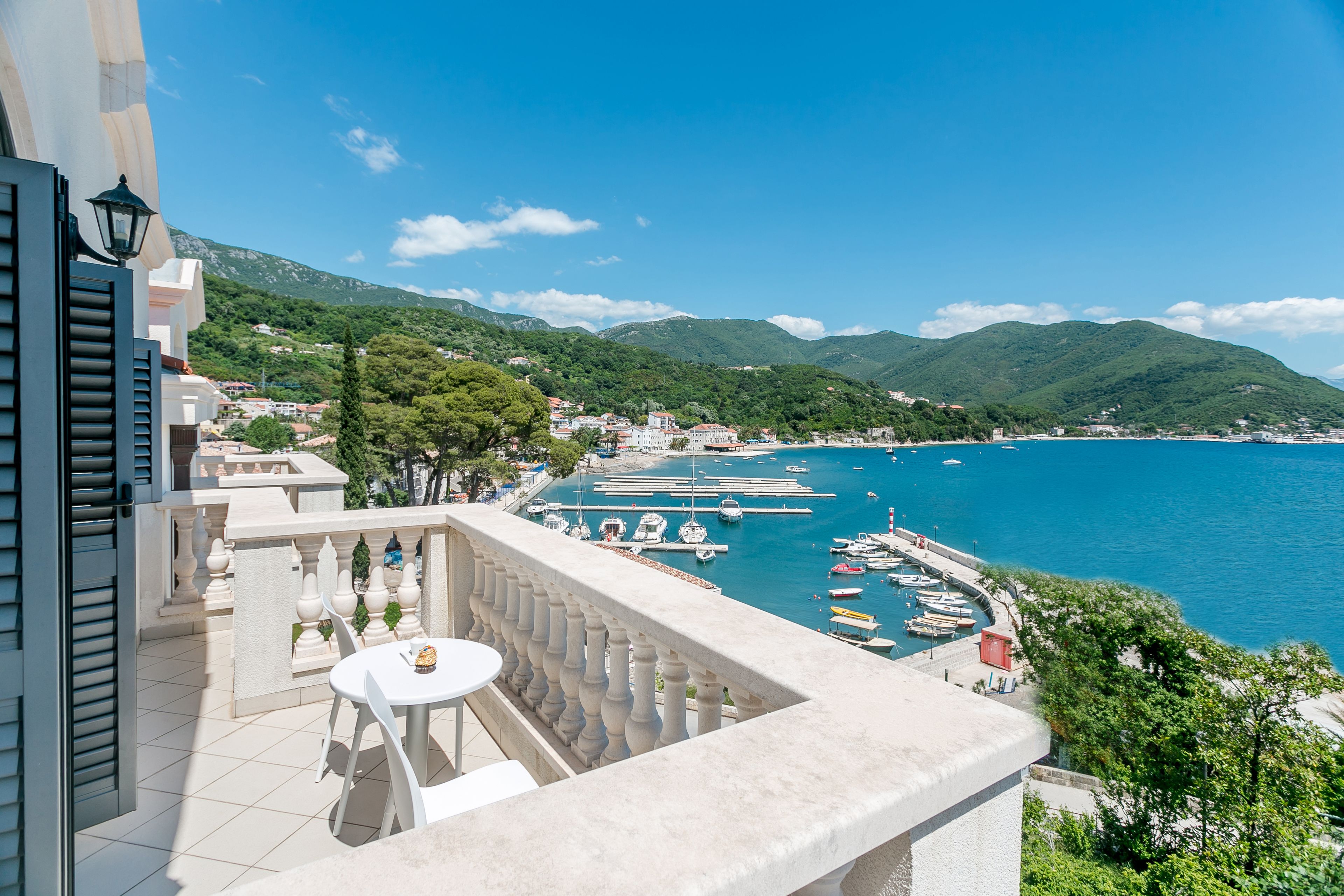 A balcony with a table and chairs overlooking a marina with boats and a mountainous landscape.