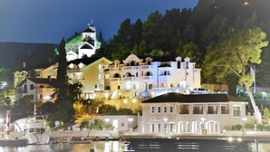 Um hotel à beira-mar com vários edifícios, incluindo uma igreja em destaque com cúpula e campanário, e um cais bem iluminado com barcos.