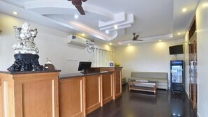 A reception area with a wooden reception desk, a statue, a ceiling fan, a television, and a seating area with a sofa and a bench. - JK Residency (Tiruchirappalli)