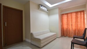A room with a sofa, a chair, an air conditioning unit, a door, and a window with curtains. - JK Residency (Tiruchirappalli)