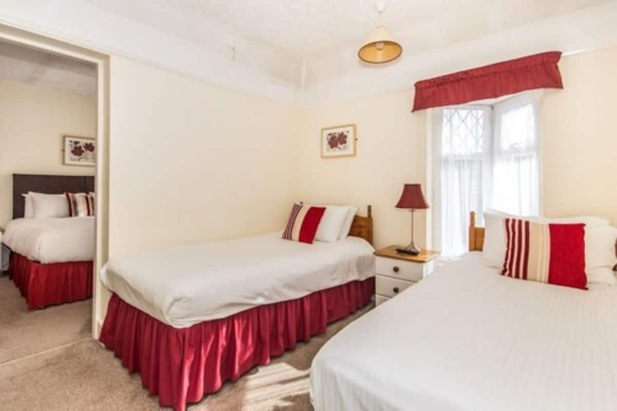 Quarto de hotel com duas camas, uma mesa de cabeceira, uma luminária e uma janela com cortinas.