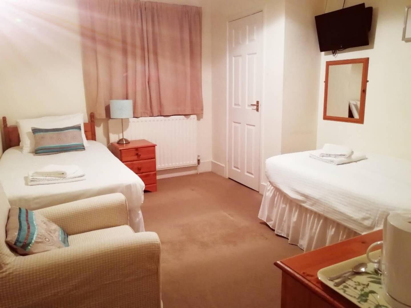 A hotel room with two beds, a small table with a cup and spoon, a chair, a nightstand, a lamp, a TV, and a door.