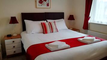 Un lit bien fait, avec une literie blanche et rouge, deux tables de chevet avec des lampes, une œuvre d’art florale encadrée accrochée au mur et une fenêtre ornée de rideaux rouges.