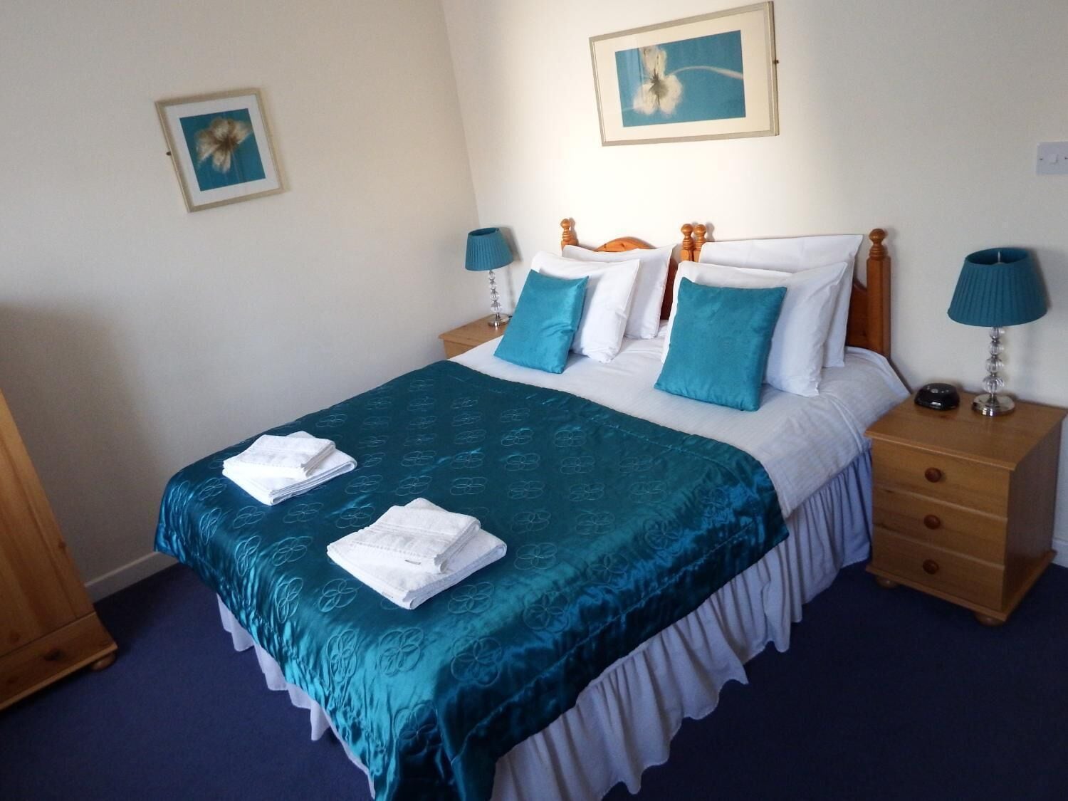 A neatly made bed with a teal bedspread and white pillows, two bedside tables with lamps, and a framed picture on the wall.