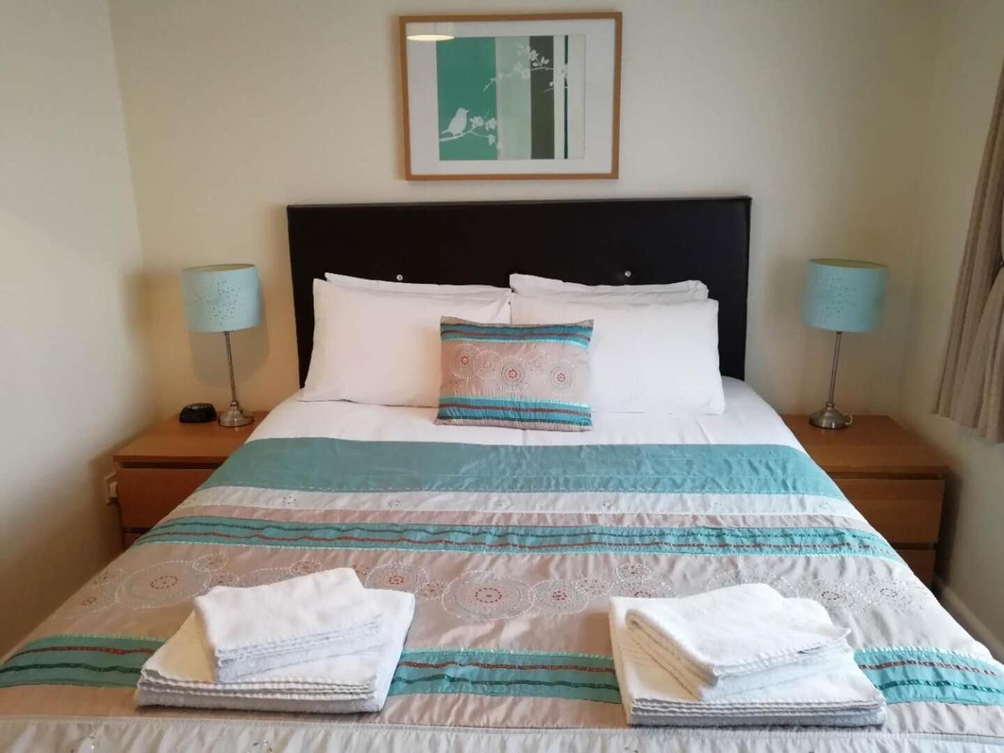 A neatly made bed with a striped bedspread, two pillows, and folded towels on a wooden nightstand.