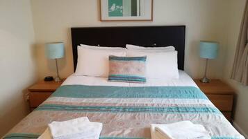 A neatly made bed with a striped bedspread, two pillows, and folded towels on a wooden nightstand.