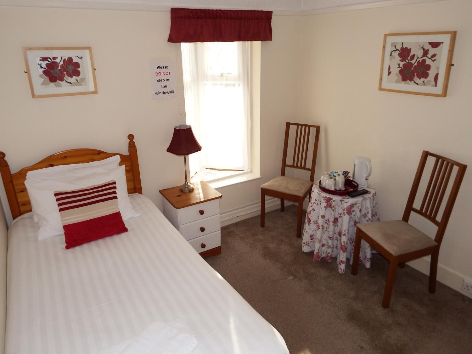 Une chambre à coucher comprenant un lit, une table de chevet, une chaise, une petite table recouverte d’une nappe à motifs floraux et une fenêtre agrémentée d’une lambrequin rouge.