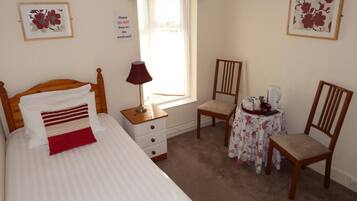 Une chambre à coucher comprenant un lit, une table de chevet, une chaise, une petite table recouverte d’une nappe à motifs floraux et une fenêtre agrémentée d’une lambrequin rouge.