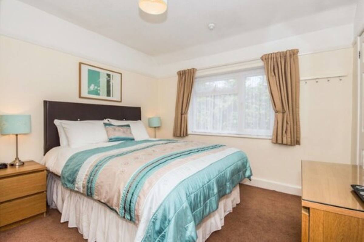 Um quarto com cama, mesas de cabeceira, uma janela com cortinas e um quadro na parede.