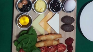 Desayuno con huevos, queso y diversas salsas sobre una tabla de madera, junto con un plato de verduras y una sartén con huevos.