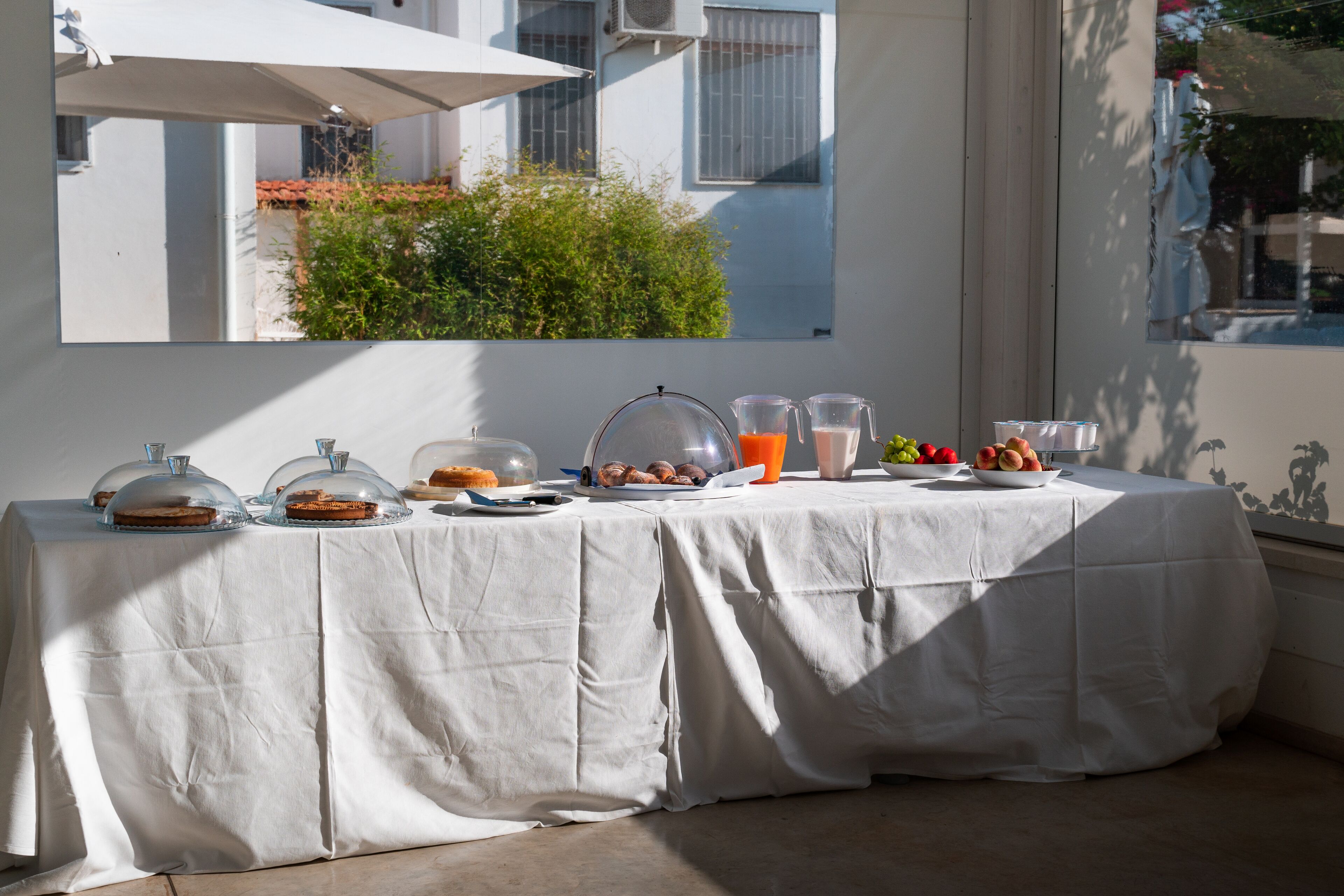 Mesa com comida e bebidas, toalha branca e vista para um edifício e vegetação lá fora.