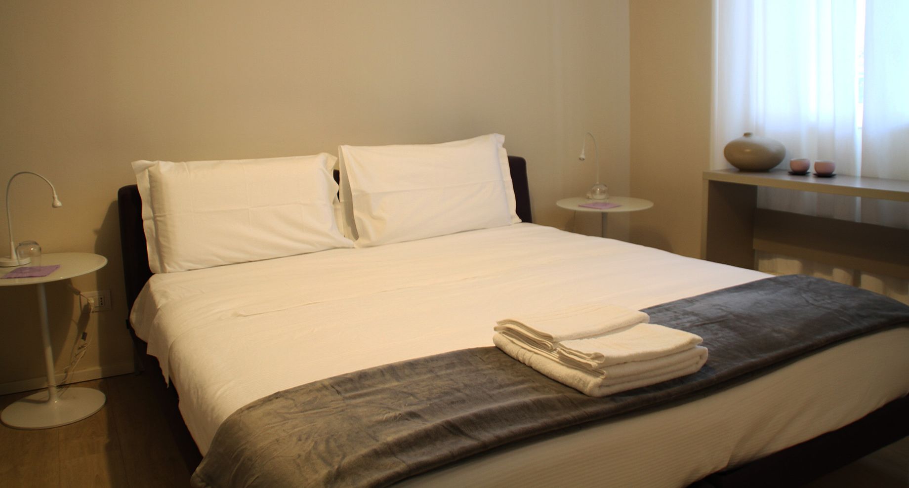 Un lit bien fait, avec du linge de lit blanc, un plaid gris et une serviette pliée posée sur le pied de lit. On y trouve une table de chevet avec une lampe et un petit vase sur la table de nuit.