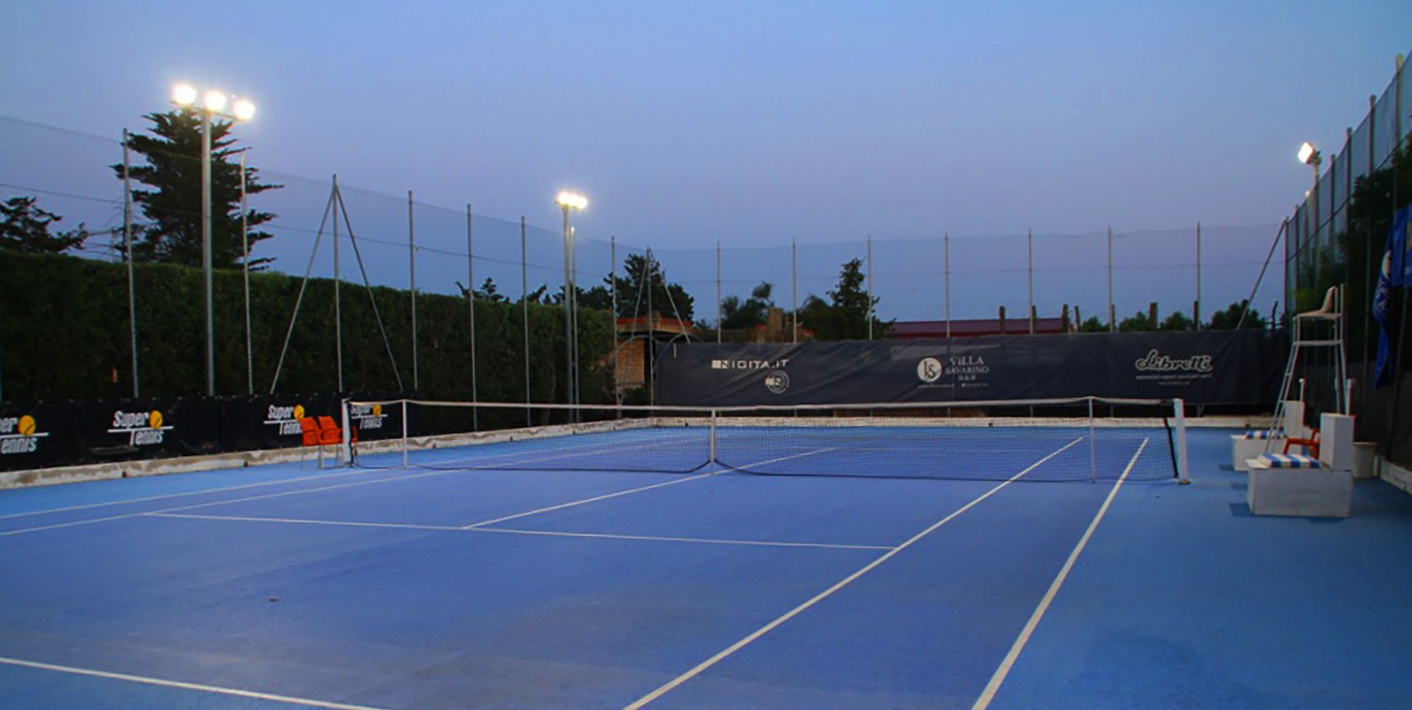 An indoor tennis court with blue flooring, net, and surrounding fencing.
