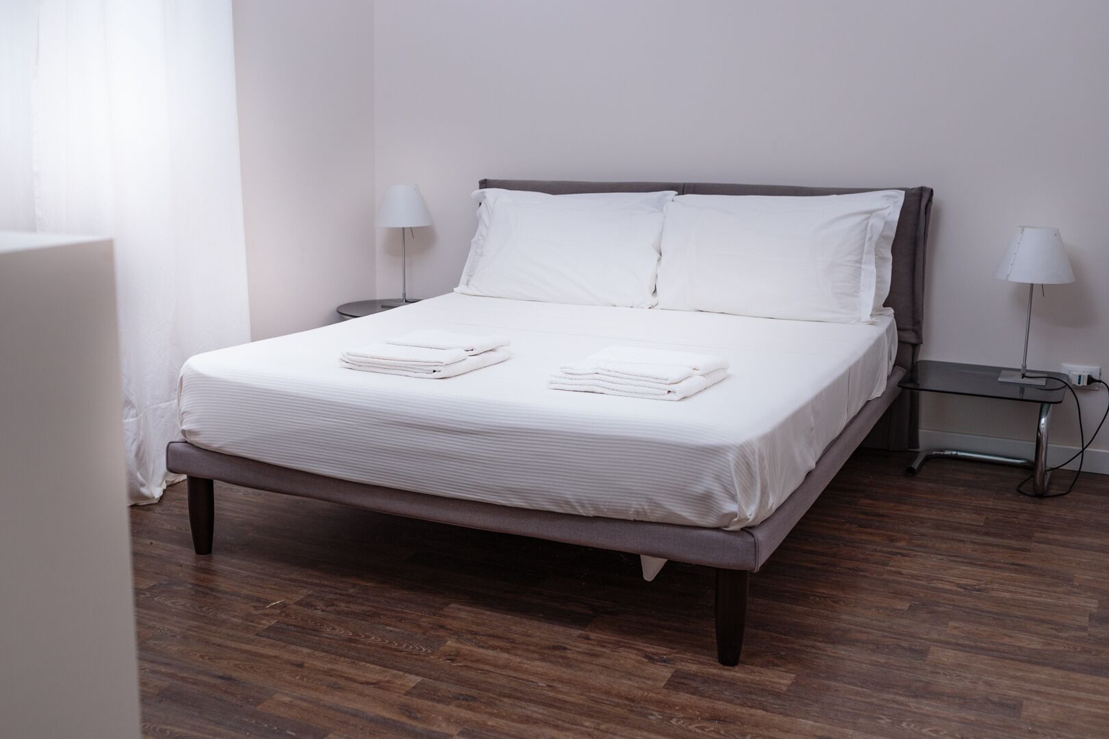 Una cama bien hecha con ropa de cama blanca y toallas dobladas, una mesita de noche con lámpara y suelo de madera.