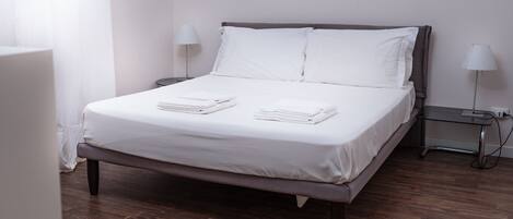 Una cama bien hecha con ropa de cama blanca y toallas dobladas, una mesita de noche con lámpara y suelo de madera.