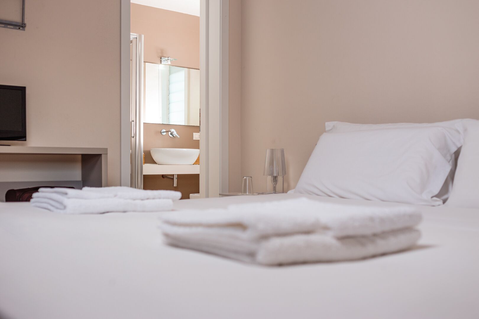 Un lit bien fait, avec du linge de lit blanc et des oreillers, une table de chevet avec une lampe, et une vue sur une salle de bain équipée d’un lavabo et d’un miroir.
