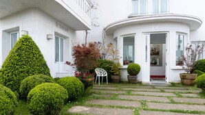 Un bâtiment blanc abritant un restaurant et un hôtel. Devant, on trouve un jardin bien entretenu, avec des arbustes taillés et une chaise.