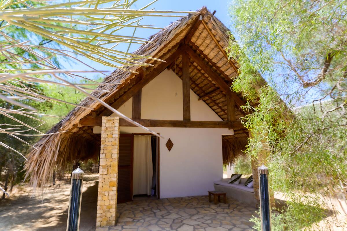 A traditional thatched-roof hut with a stone foundation, a wooden door, and a small bench outside.