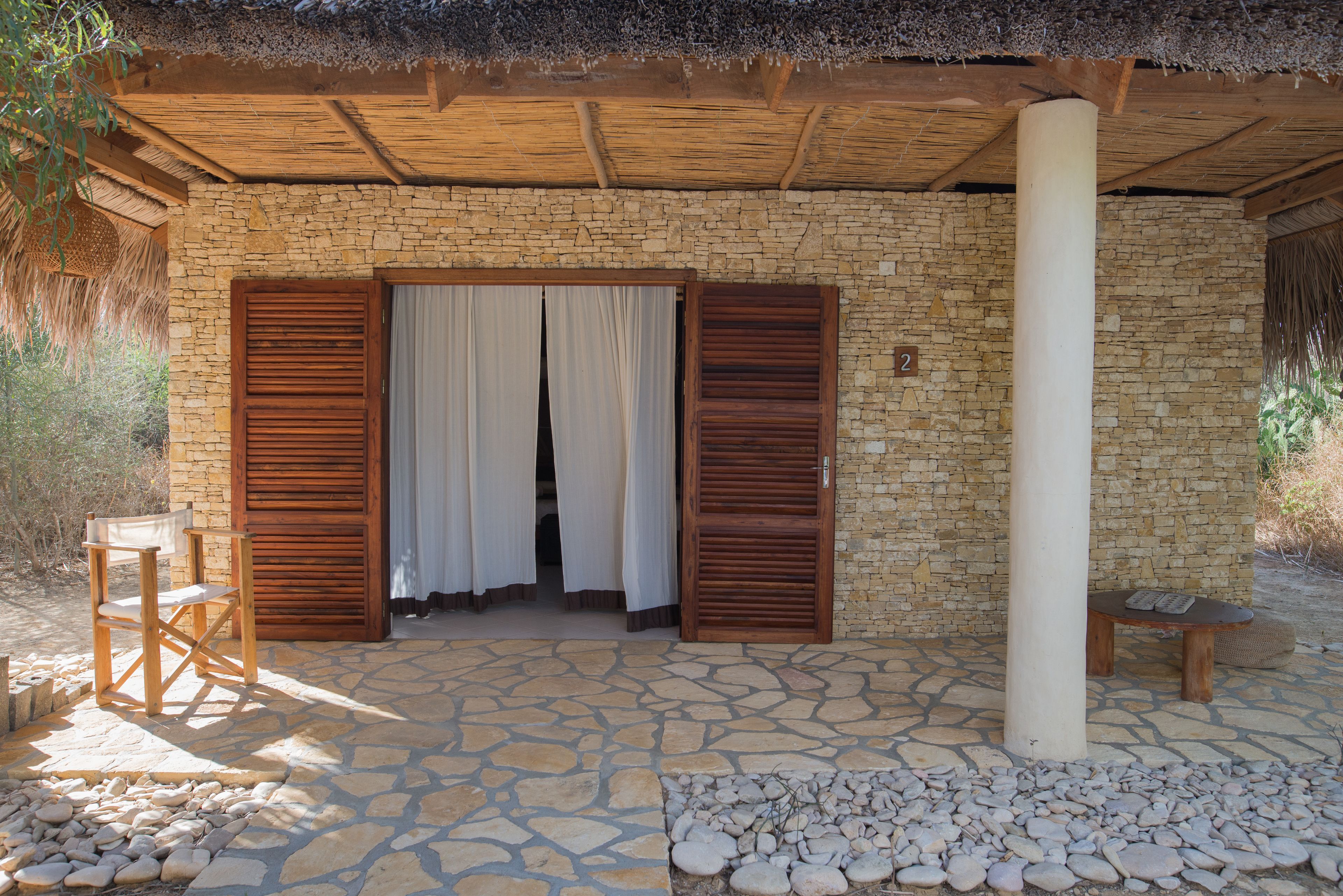 A stone-walled room with wooden shutters, a thatched roof, and a stone floor.