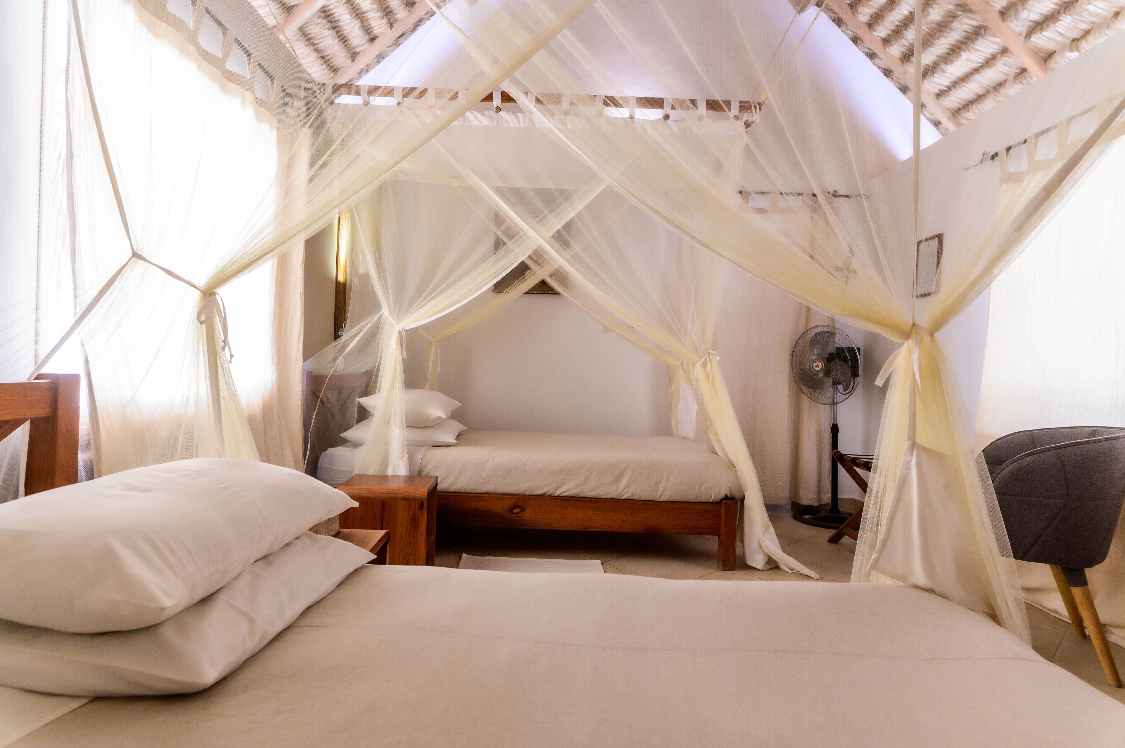 A bedroom with a canopy bed, a bedside table, a chair, and a standing fan.
