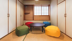 Una habitación japonesa tradicional con suelo de tatami, ventana con pantalla shoji y una mesa baja de tatami rodeada de cojines coloridos.