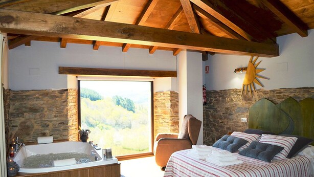 Une chambre avec un mur en pierre, un plafond en bois, une grande fenêtre, une baignoire et un lit avec des oreillers.
