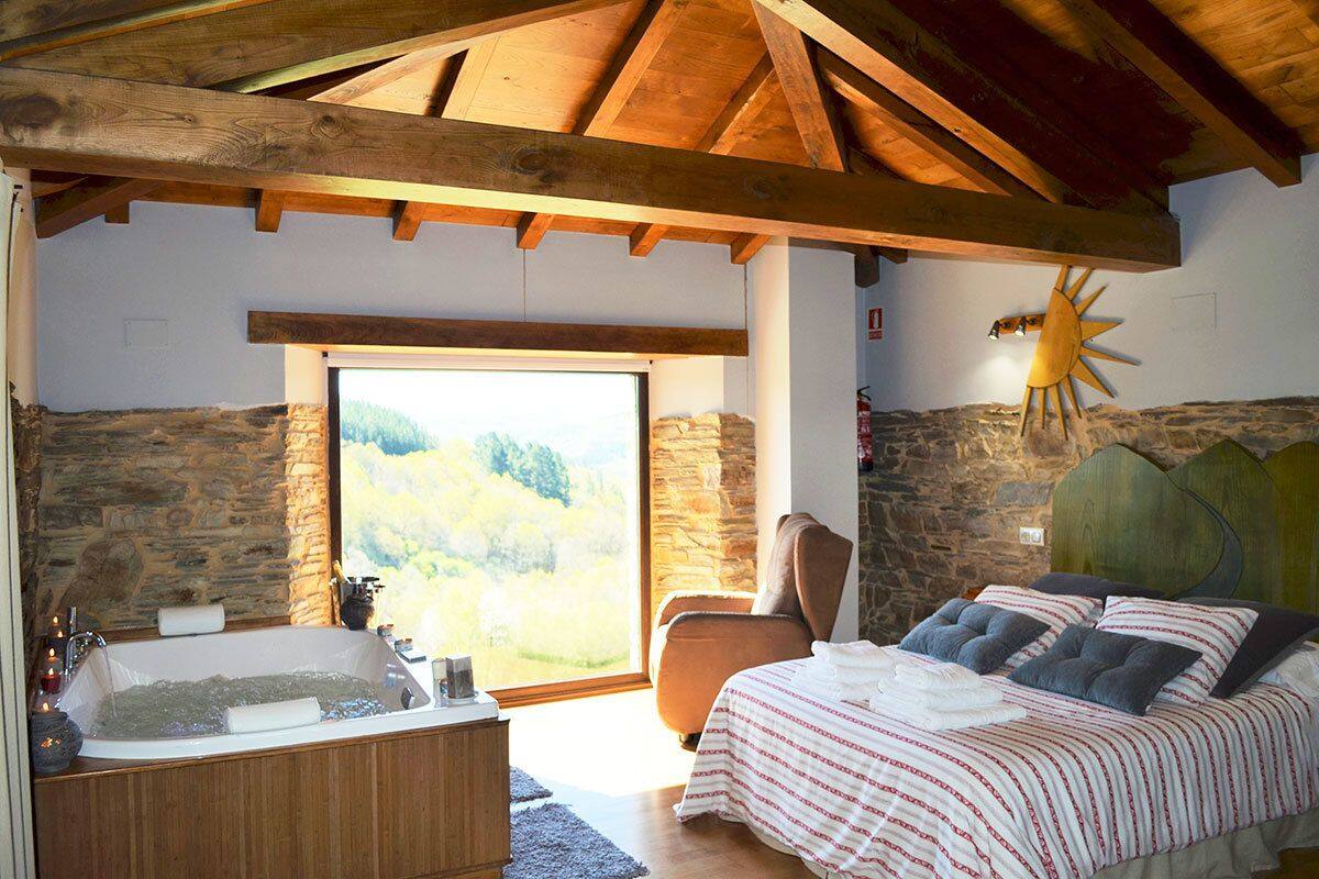 A bedroom with a stone wall, a wooden ceiling, a large window, a bathtub, and a bed with pillows.