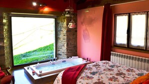 Une pièce avec un mur en pierre, une grande fenêtre donnant sur un paysage verdoyant, un lit en bois avec une literie à motifs floraux et un jacuzzi.