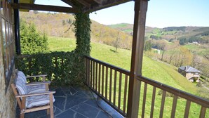 Un balcon abrité avec un sol en pierre, un banc en bois et une vue sur une vallée verdoyante, avec des arbres et une maison au loin.