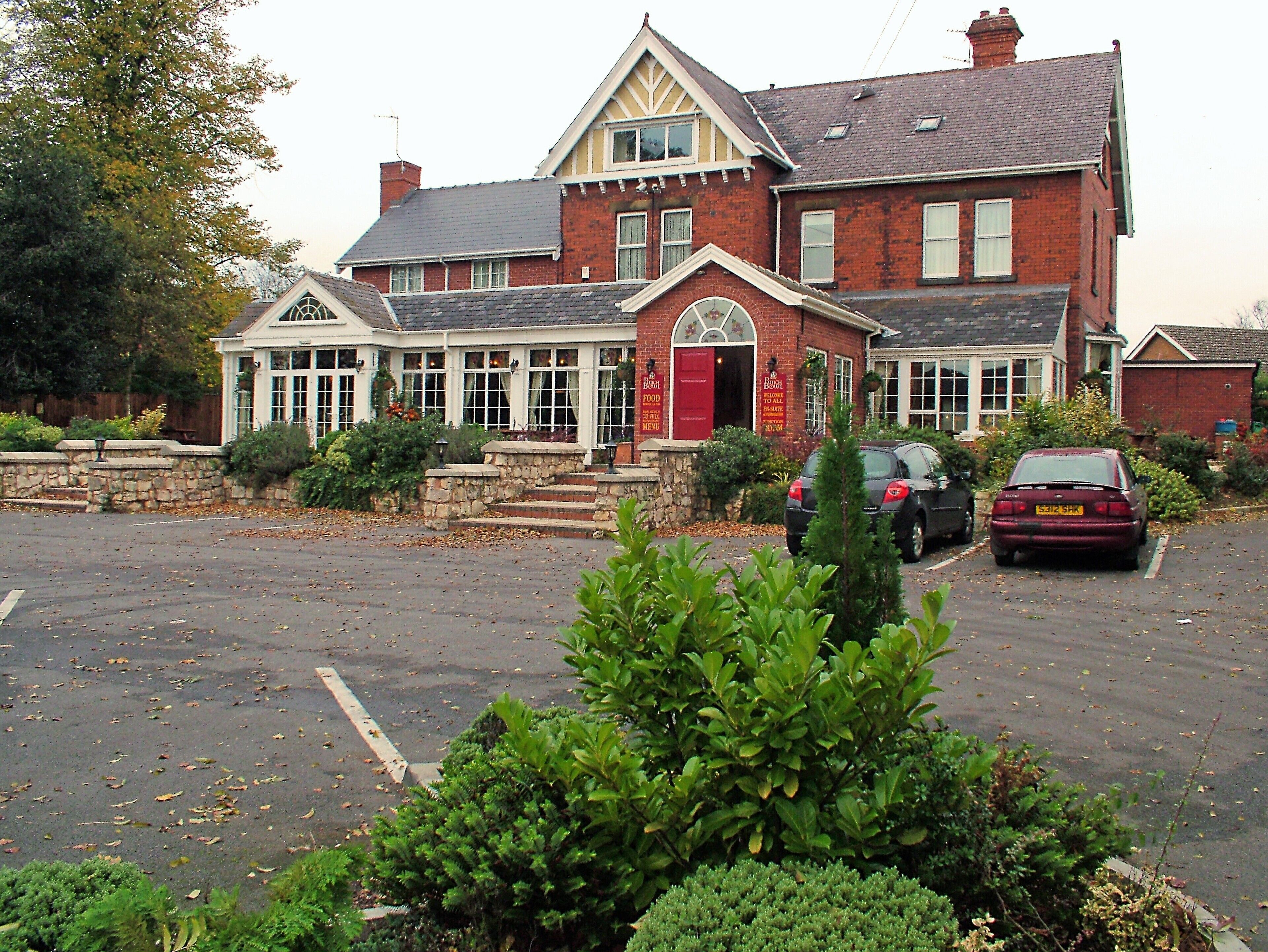 A two-story building with a red door and multiple windows, surrounded by greenery and parked cars.