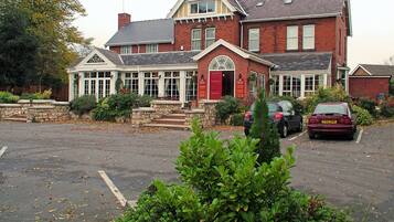 A two-story building with a red door and multiple windows, surrounded by greenery and parked cars.