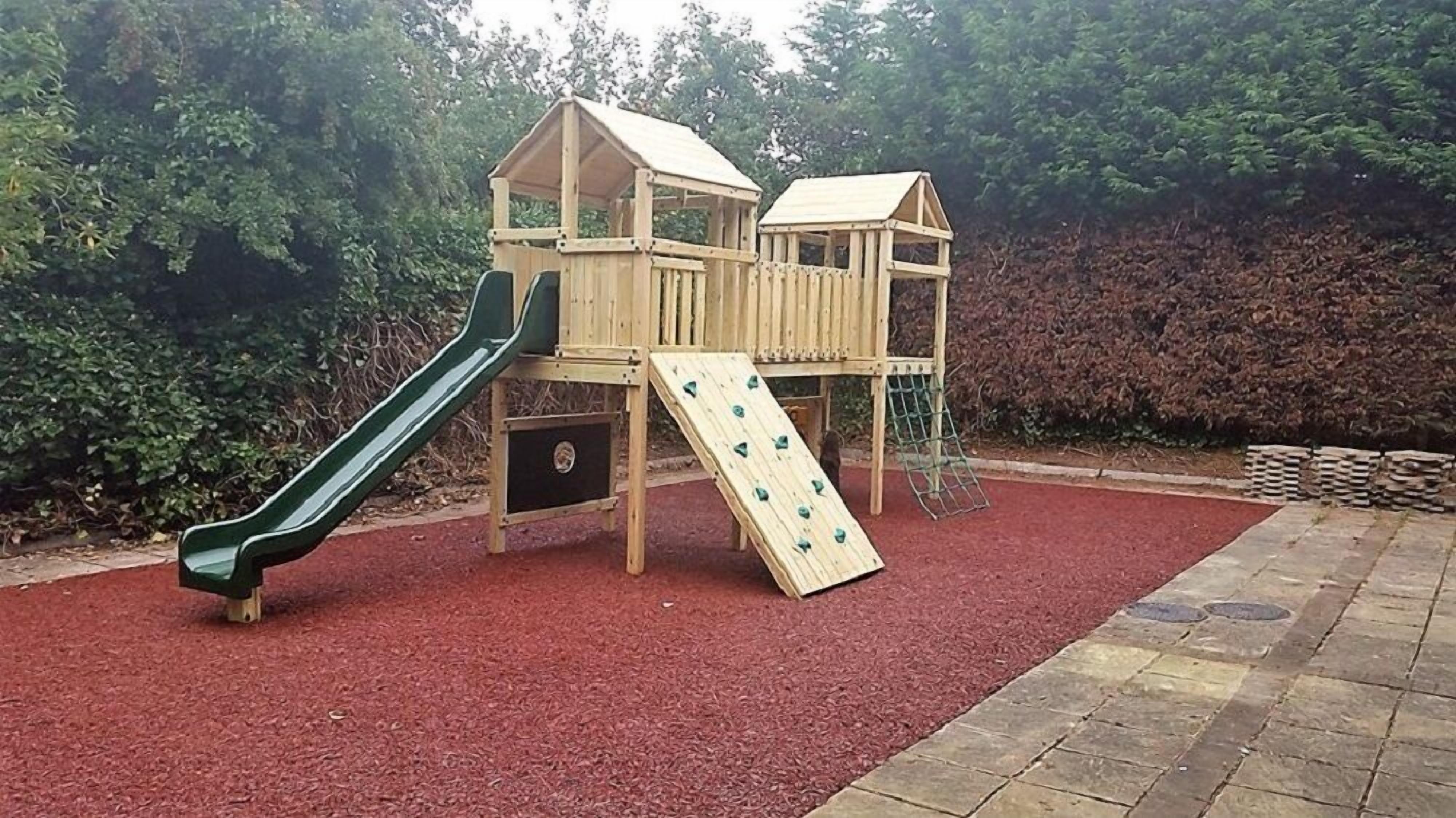 A wooden play structure with a slide and climbing wall, set on red mulch ground.